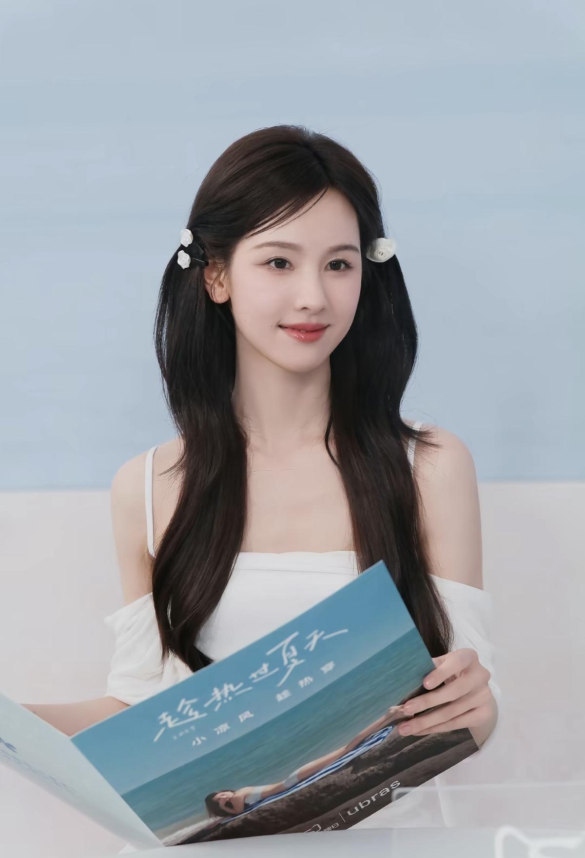 陈都灵一袭白裙配双马尾，发间的小花点缀得恰到好处，手里的夏日主题册子仿佛也带着海
